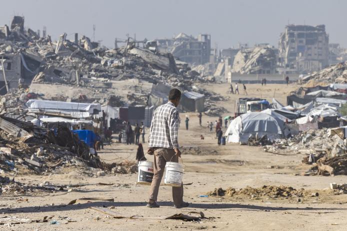 Palestinian man, holding buckets, walks among rubble of Gaza.